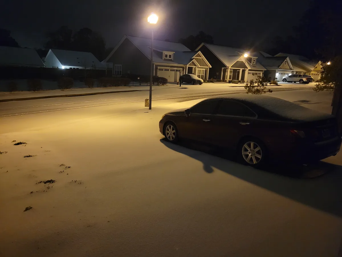 A nighttime photo of a snow-covered suburban street with houses and cars under streetlights. A dark sedan is parked on the…