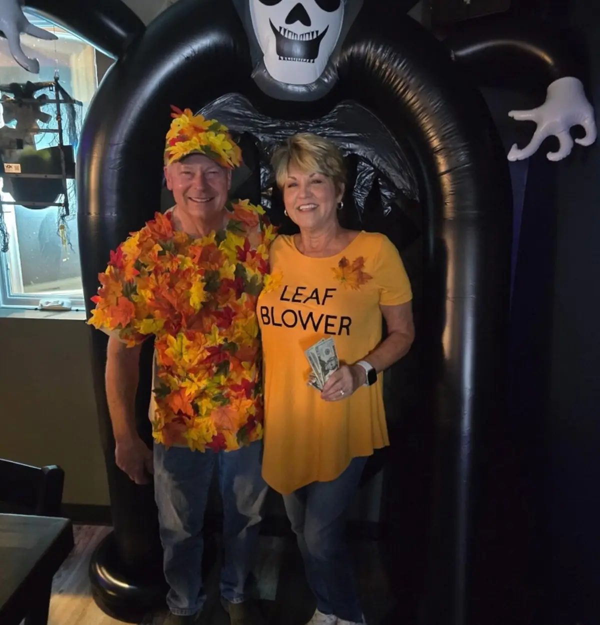 A smiling man and woman pose in front of a large inflatable skeleton archway. The man wears a shirt covered in autumn leaves…