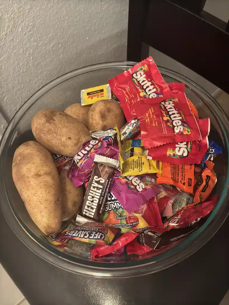 A clear glass bowl contains several unpeeled potatoes mixed with various colorful candy packages including Skittles…