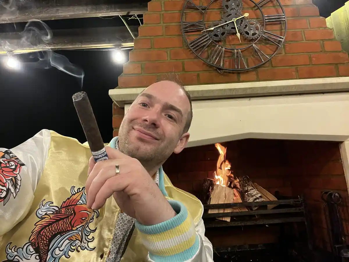 A man in a yellow satin jacket with koi fish and rose embroidery holds a lit cigar, smiling smugly at the camera. Behind…