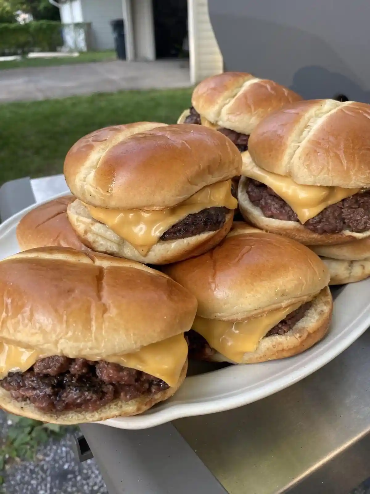 A white plate holds several cheeseburgers with golden-brown buns and melted yellow cheese, stacked and arranged outdoors on…