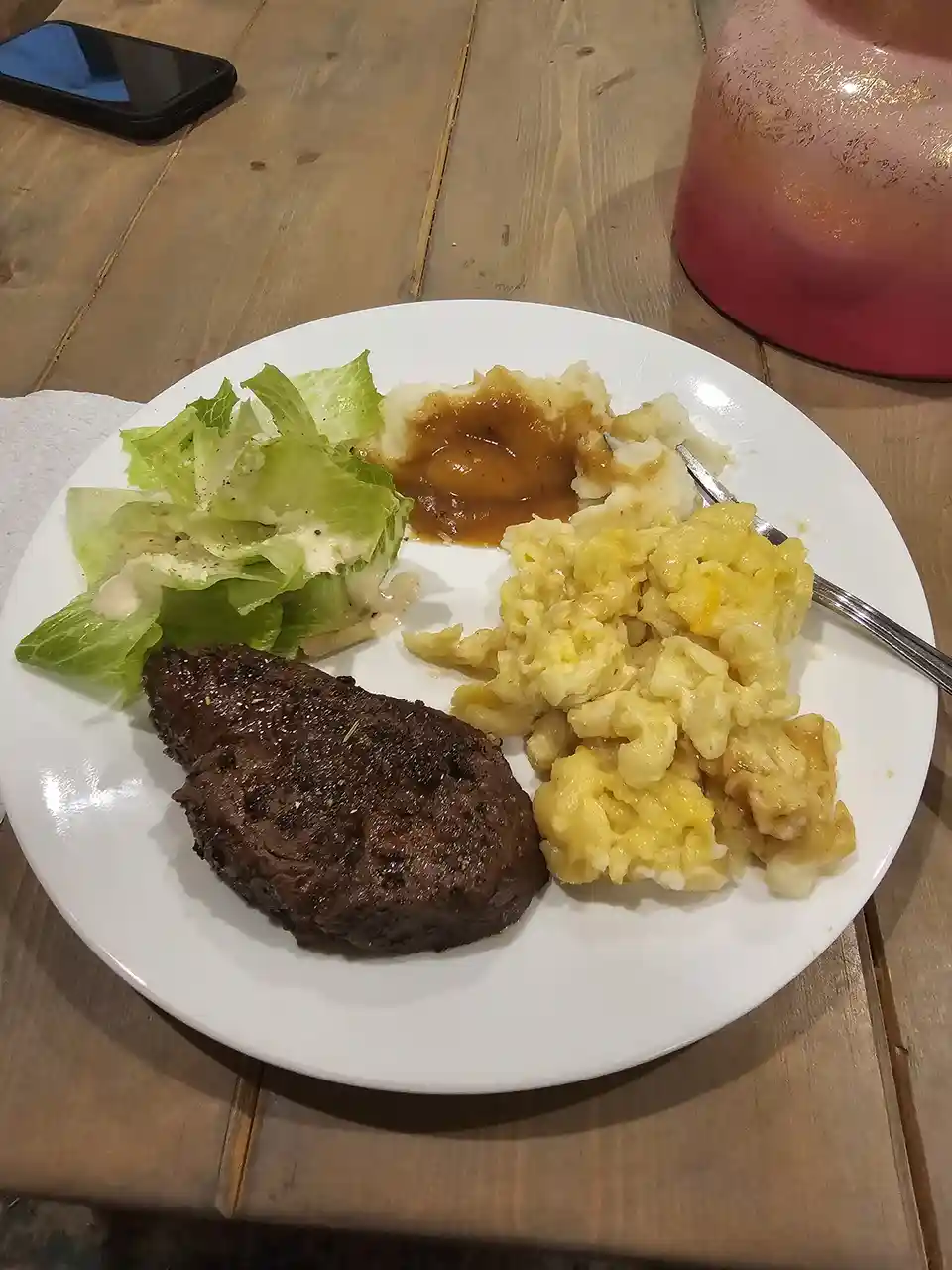 A white plate on a wooden table holds a meal including a seared meat patty, macaroni and cheese, mashed potatoes with gravy…