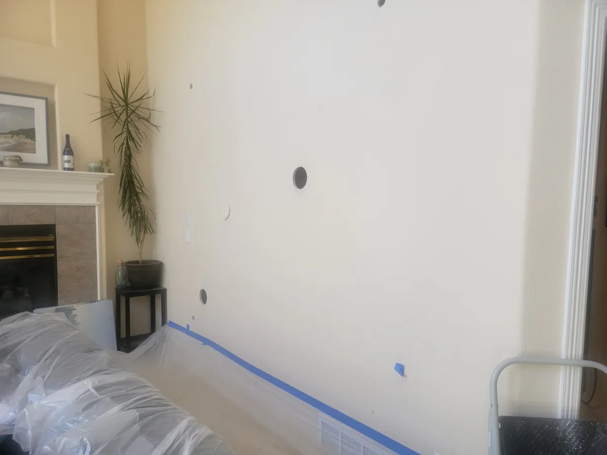 A room under renovation shows a freshly painted wall with multiple circular holes and patched areas, blue painter's tape…