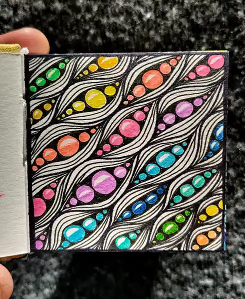 A hand holds a small, intricately drawn card featuring swirling black-and-white patterns filled with colorful…