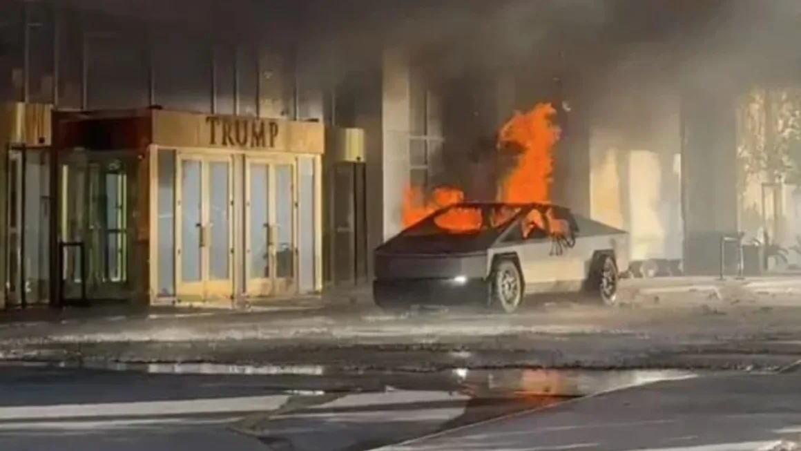 A Tesla Cybertruck is engulfed in flames on a street in front of a building with a golden entrance that reads 'TRUMP'. The…