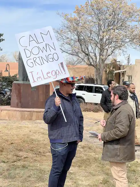 A man wearing a colorful sombrero and sunglasses holds a handmade sign that reads 'CALM DOWN GRINGO I'm Legal!' while…