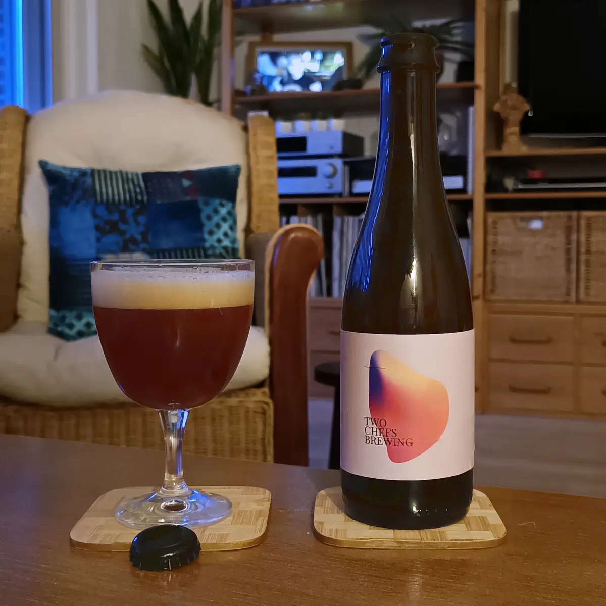 A dark amber beer in a stemmed glass sits next to its bottle labeled 'TWO CHEFS BREWING' on a wooden table, with a cozy…