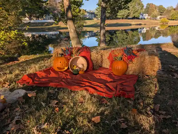 An outdoor autumn scene shows two hay bales arranged like couch cushions, covered with a red blanket and decorated with…