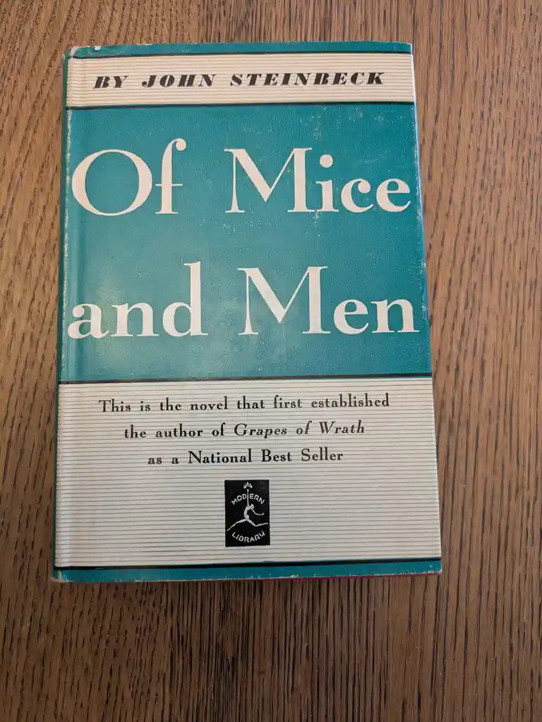 A vintage teal and cream hardcover of John Steinbeck's 'Of Mice and Men' rests on a wooden surface, with text noting it…