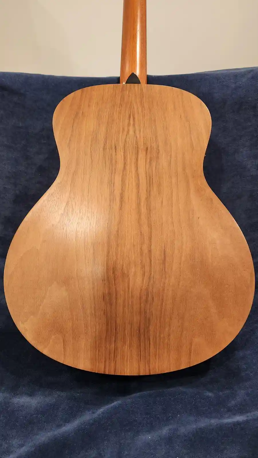 The image shows the back of an acoustic guitar with a light brown wooden body and a dark blue fabric background. The…
