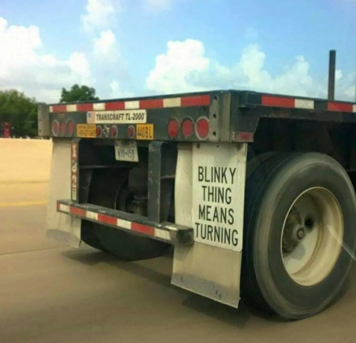 The rear of a flatbed semi-truck with red and white reflective tape, showing a Texas license plate and a sign that reads…