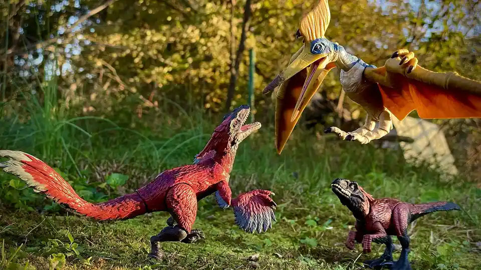 Three plastic dinosaur toys are staged outdoors in grassy terrain: a large red theropod lunges forward, a smaller dark…