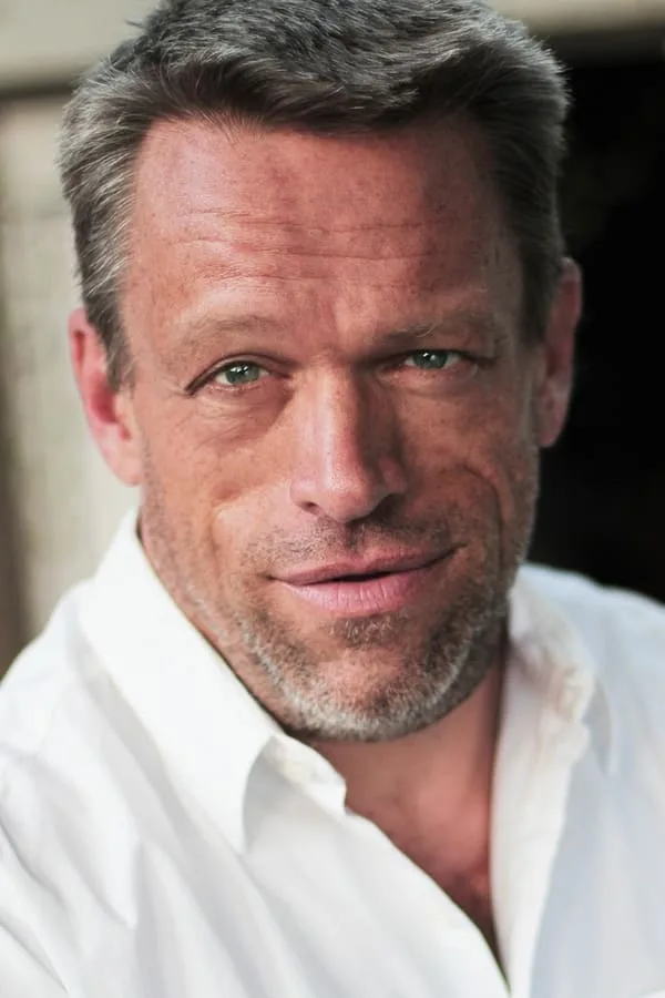 A close-up portrait of a middle-aged man with graying hair and a light beard, wearing a white collared shirt. He has a…