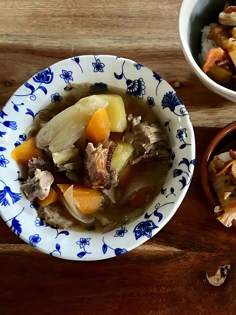 A close-up photo shows a bowl of hearty stew with chunks of meat, carrots, and potatoes in a brown broth, served in a white…