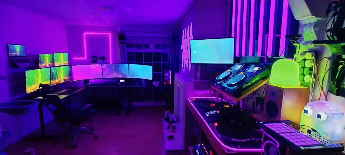 A room bathed in purple and green neon light features a large multi-monitor gaming setup on the left and a DJ station with…