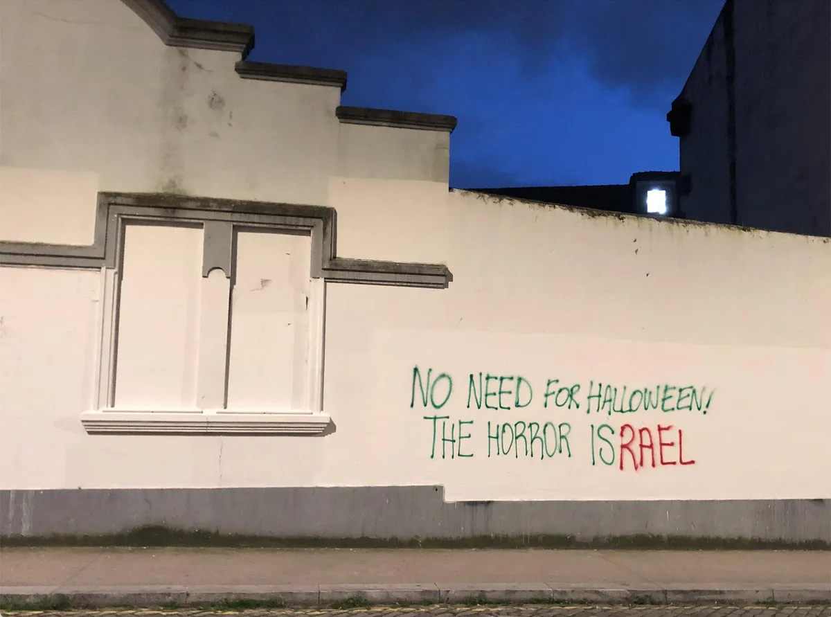 A nighttime photo of a white building wall with a window, showing green and red spray-painted text that reads 'NO NEED FOR…