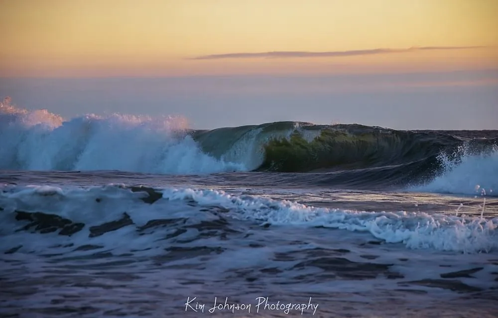 A serene ocean scene at sunset, featuring a large, curling wave with white foam crashing against a calm foreground. The sky…