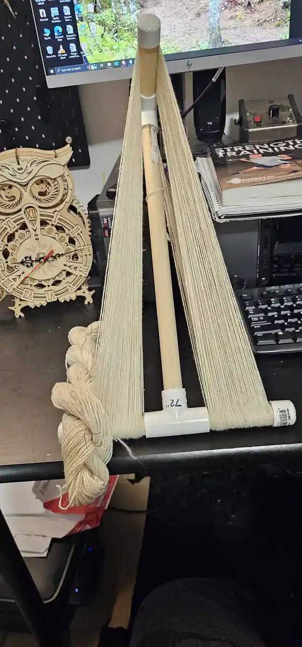 A DIY yarn swift made from a wooden baseball bat and white PVC pipes sits on a desk, holding numerous strands of beige yarn…