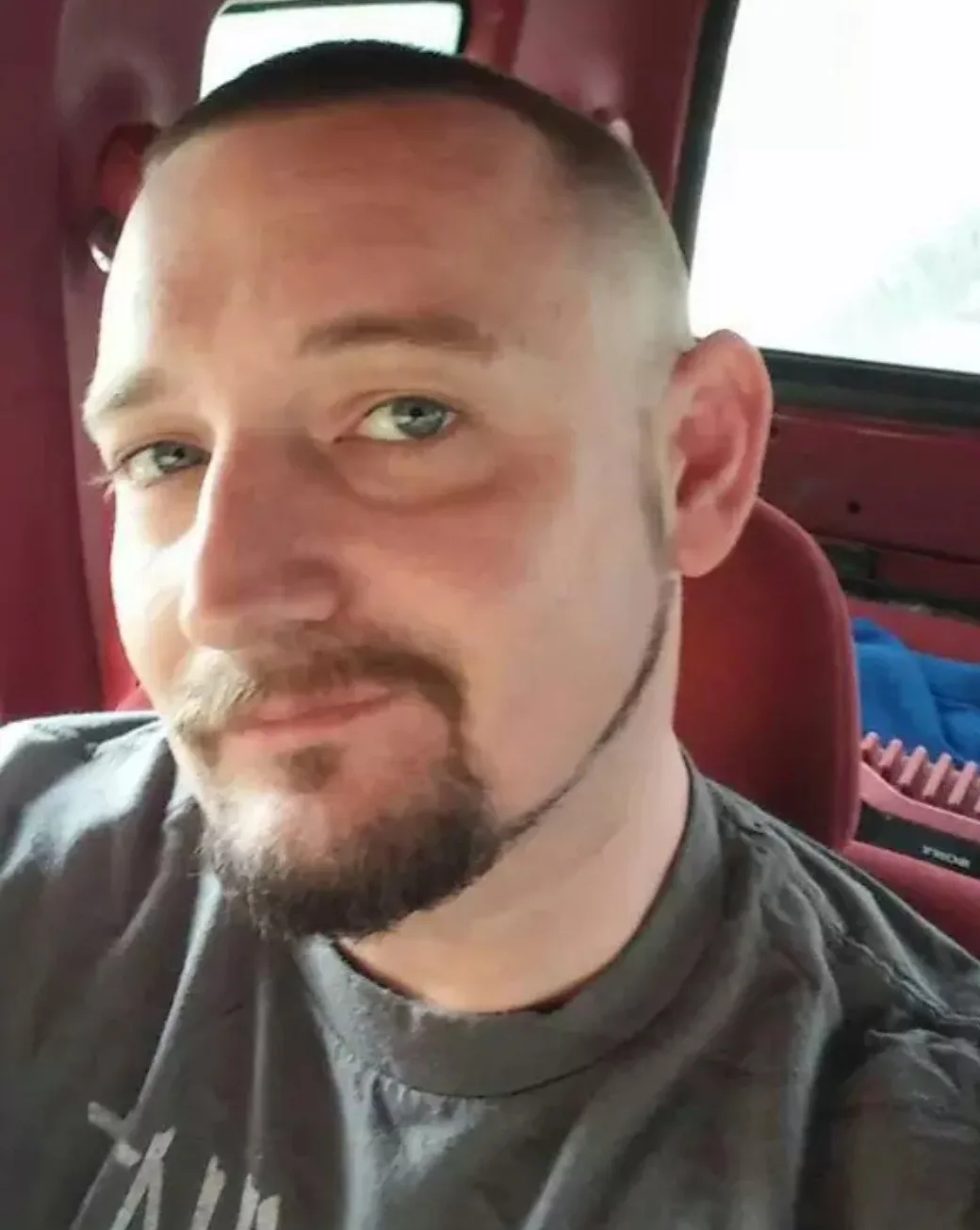 A man with a very short buzz cut and a goatee is sitting in a red car seat, looking at the camera with a slight smirk. The…