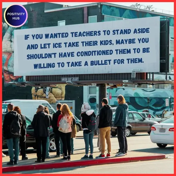 A large billboard displays a sarcastic message about teachers and ICE, while a group of people stand on a sidewalk below it…