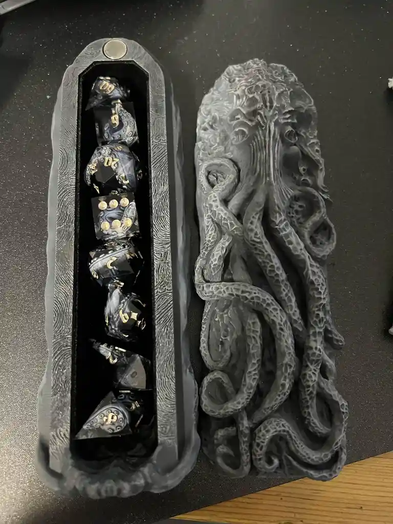 A dark grey, textured dice tray shaped like Cthulhu's head and tentacles holds a set of black dice with gold numbers. The…