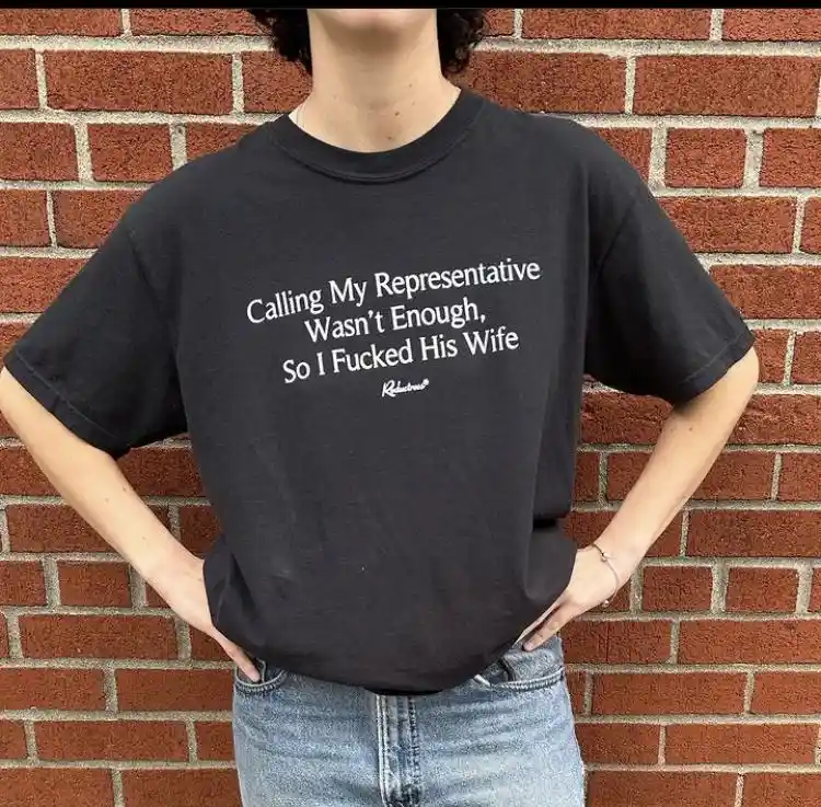 A person wearing a black t-shirt with white text that reads 'Calling My Representative Wasn't Enough, So I Fucked His Wife'…