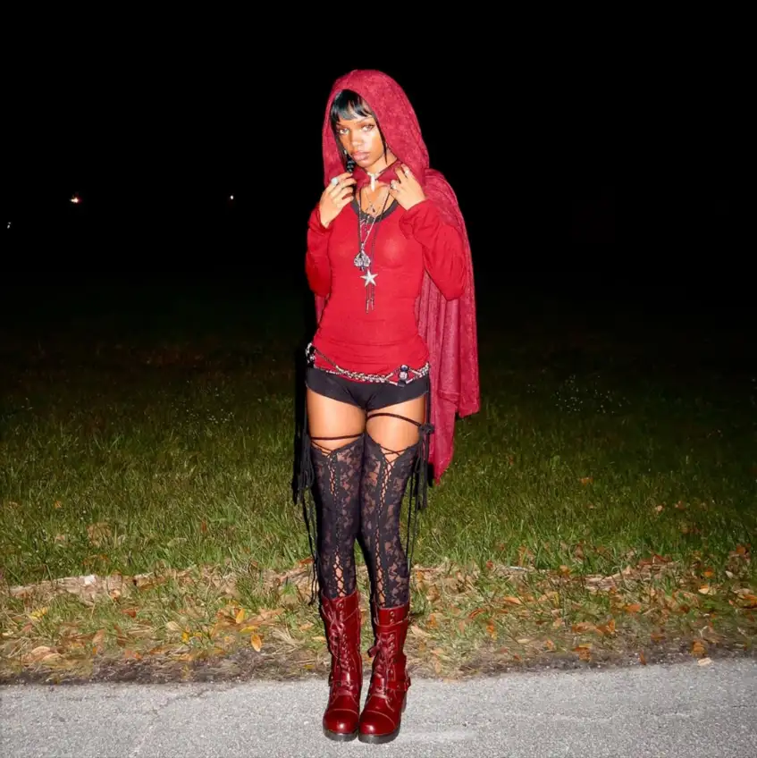 A person stands outdoors at night wearing a red hooded outfit, black shorts, lace thigh-high stockings, and red boots, with…