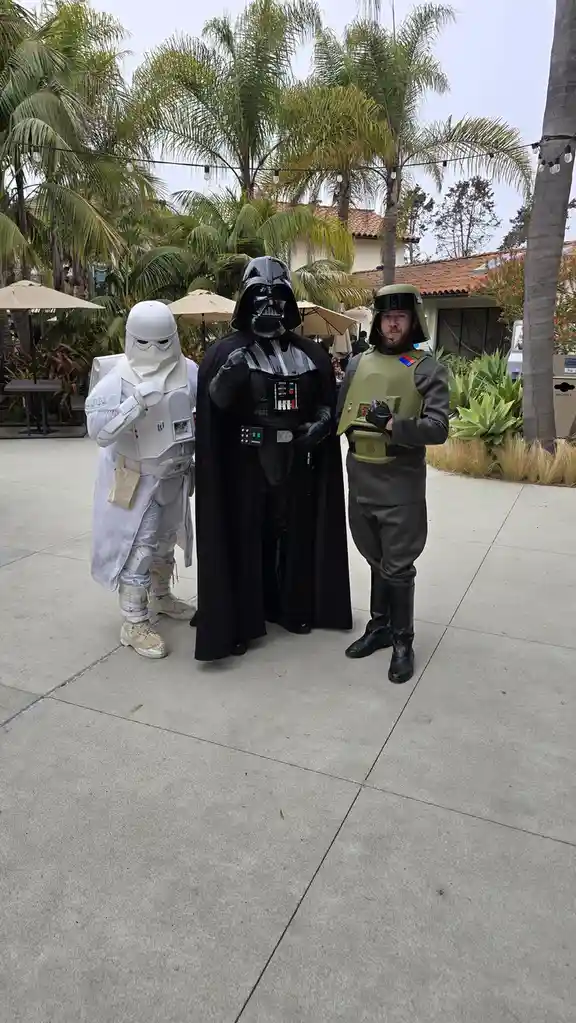 Three people dressed as Star Wars characters stand side-by-side outdoors: a white-armored stormtrooper on the left, Darth…