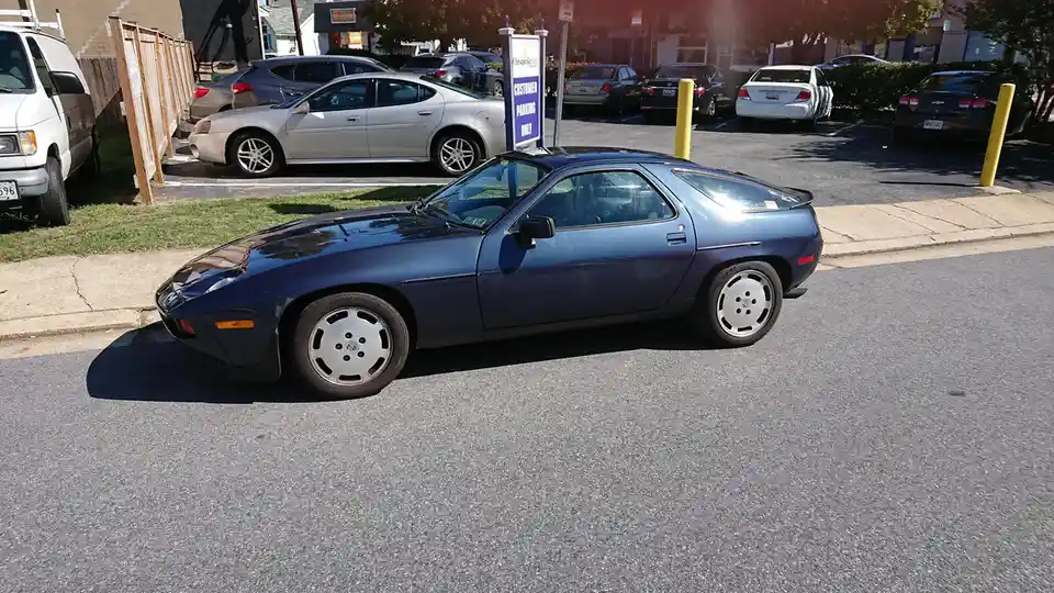 A dark blue Porsche 924 parked on a street next to a parking lot with a sign that reads 'CUSTOMER PARKING ONLY'. The car is…