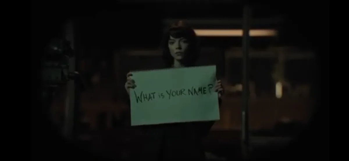 A woman with dark hair stands in a dimly lit, blurry background holding a green spiral-bound notebook with handwritten text…