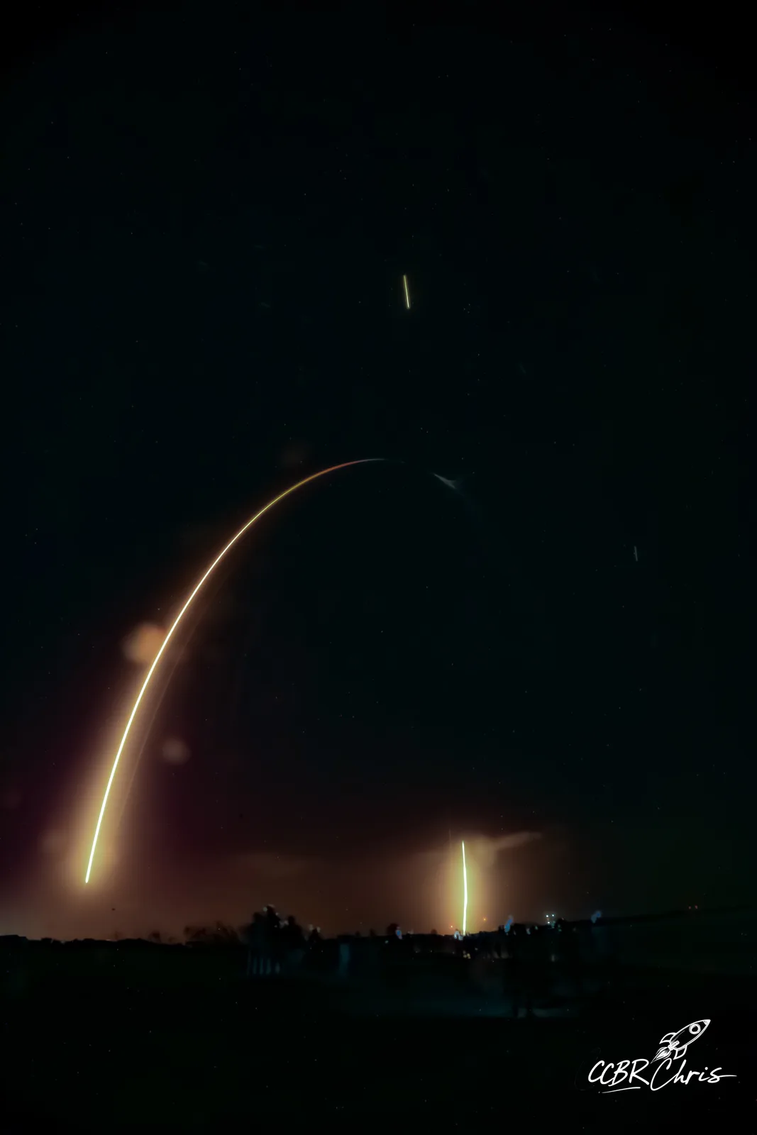 A long-exposure photograph captures a bright, curved light trail of a rocket ascending into a starry night sky, with…