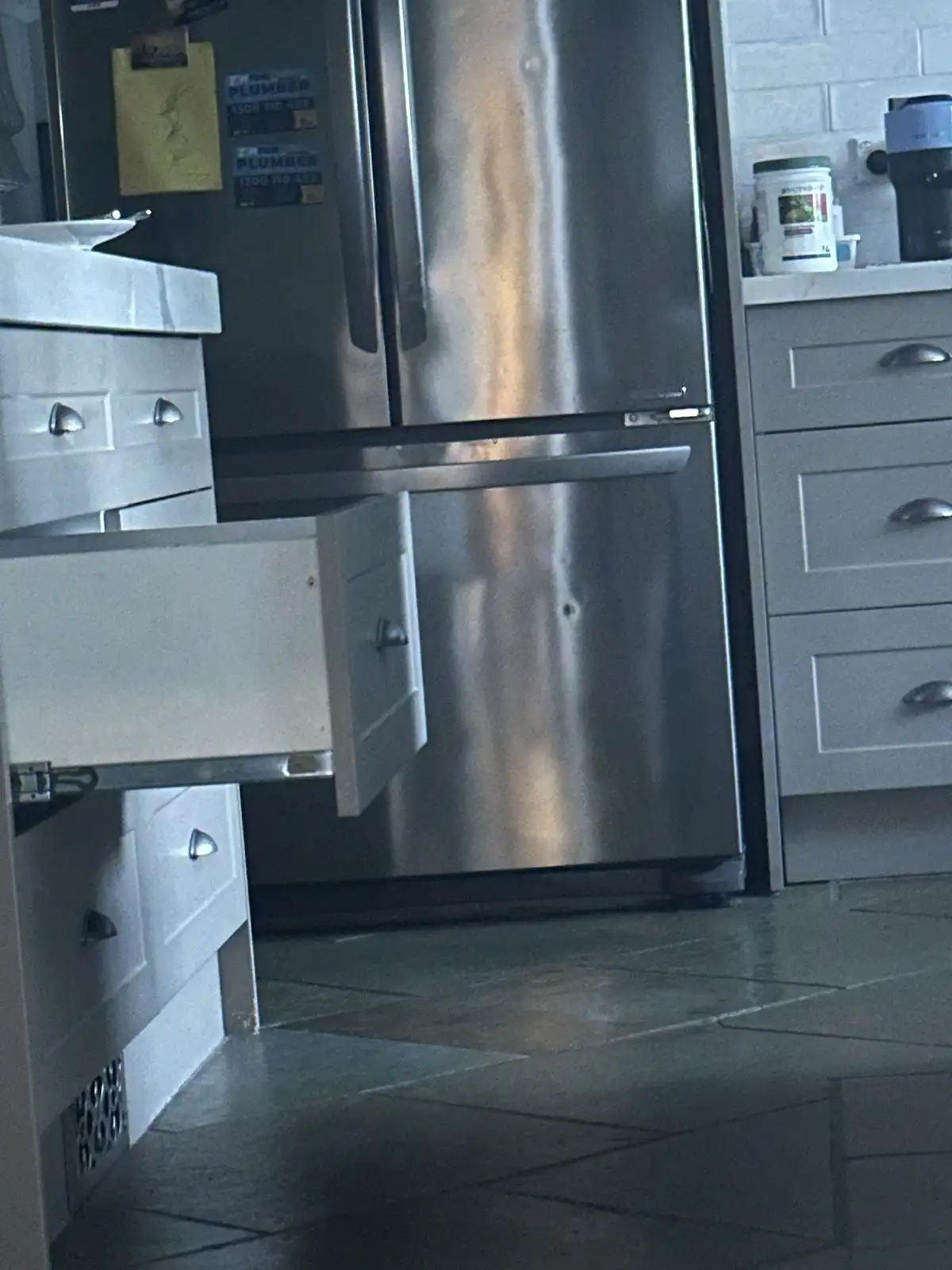 A stainless steel refrigerator stands in a kitchen with light gray cabinets; a drawer is pulled open to the left, revealing…