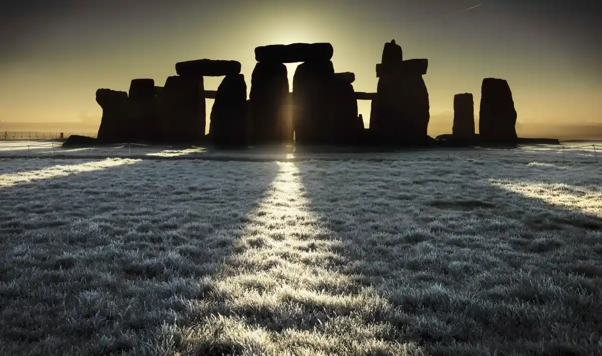 A serene, frost-covered field leads to the silhouette of Stonehenge at sunrise, with the sun casting a bright path through…