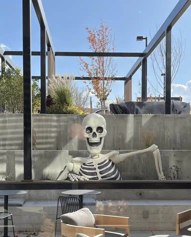 A large, white plastic skeleton decoration is lounging casually on a concrete ledge in an outdoor patio area, propped up…