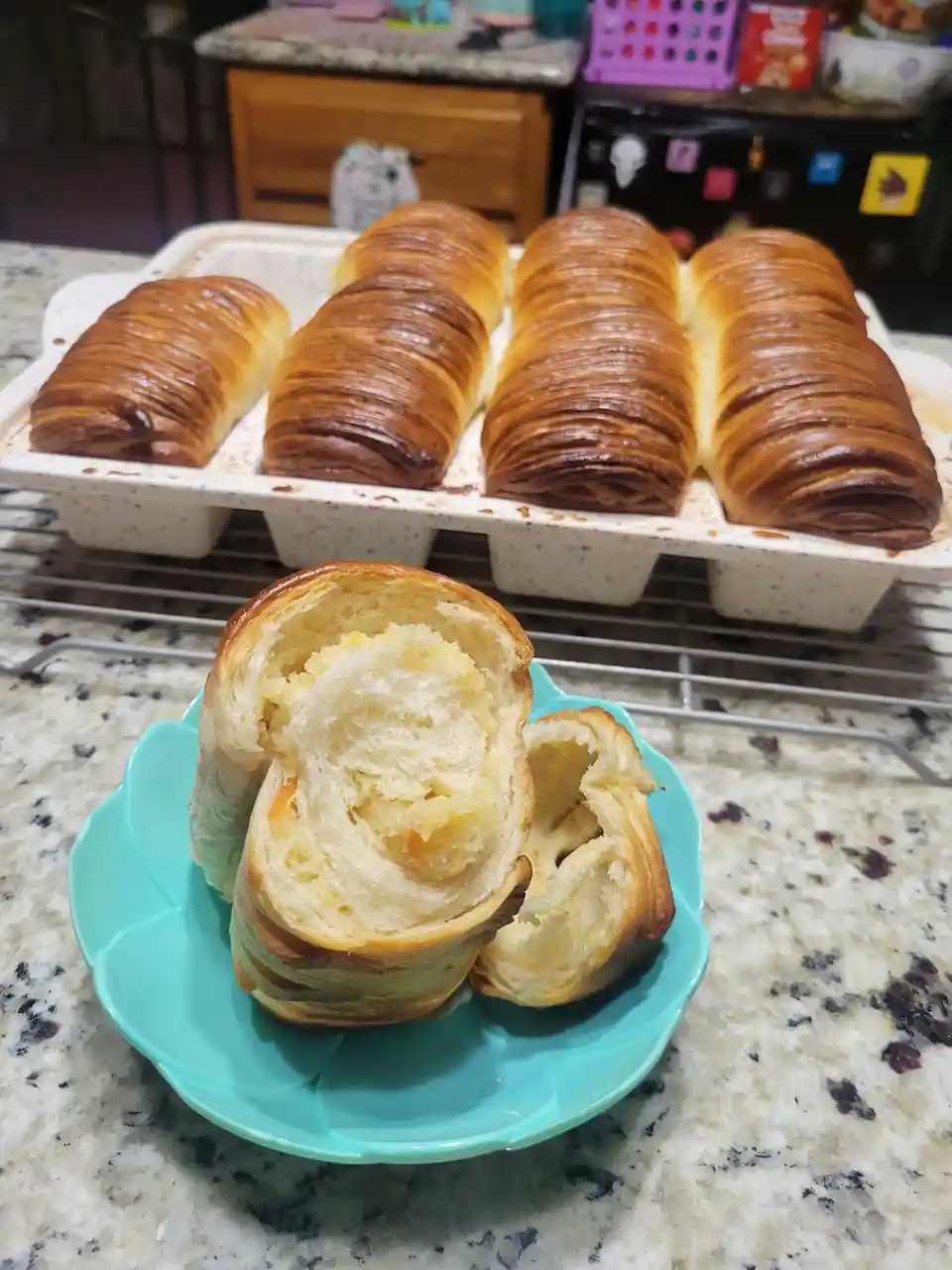 A kitchen scene shows four golden-brown croissants in a white speckled baking pan on a wire rack, with one croissant sliced…