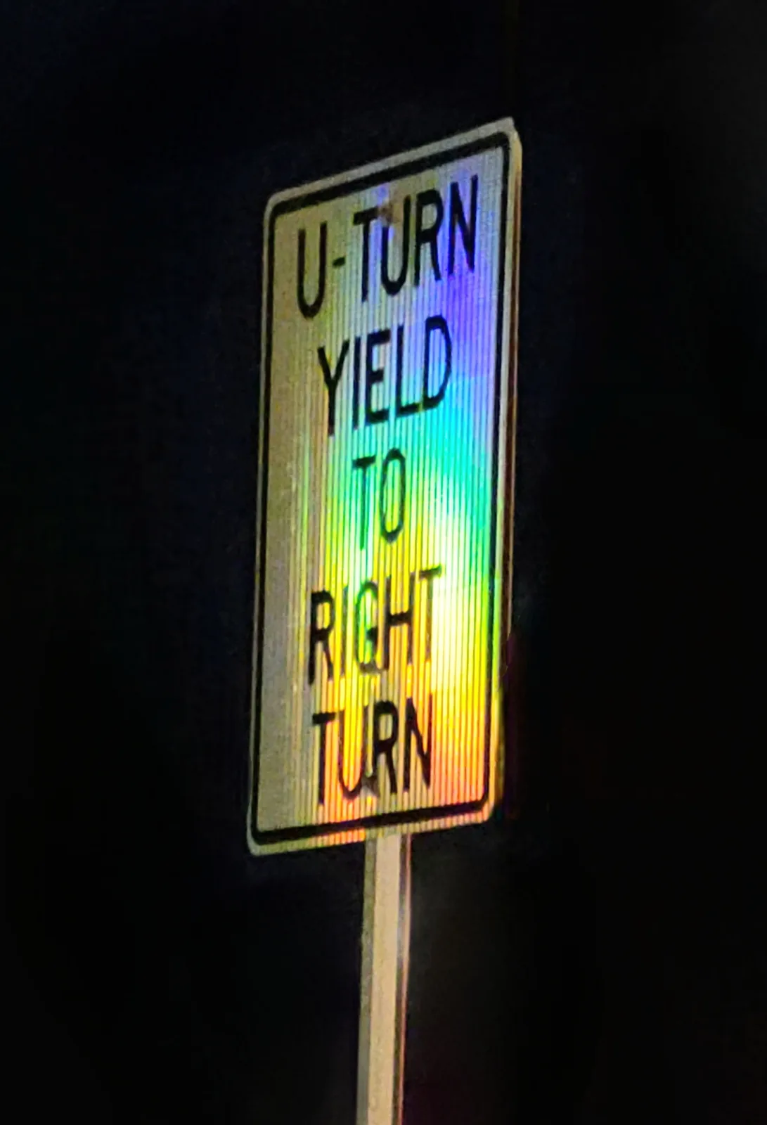 A nighttime photo of a rectangular road sign with black text reading 'U-TURN YIELD TO RIGHT TURN' against a reflective…