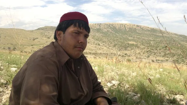 a young man wearing a red cap and brown shirt sits in a grassy field with hills in the background, looking directly at the…