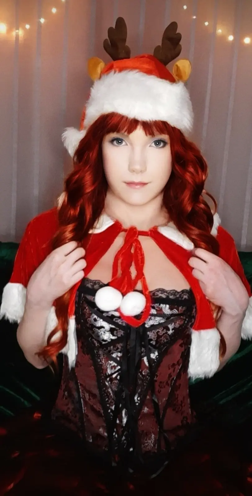 A person with red hair wears a red and white Santa hat with brown reindeer antlers and yellow ears, pulling open a red…