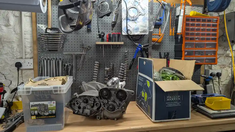 A cluttered workshop bench holds a disassembled engine block next to a large open cardboard box labeled 'PHILIP WARREN…