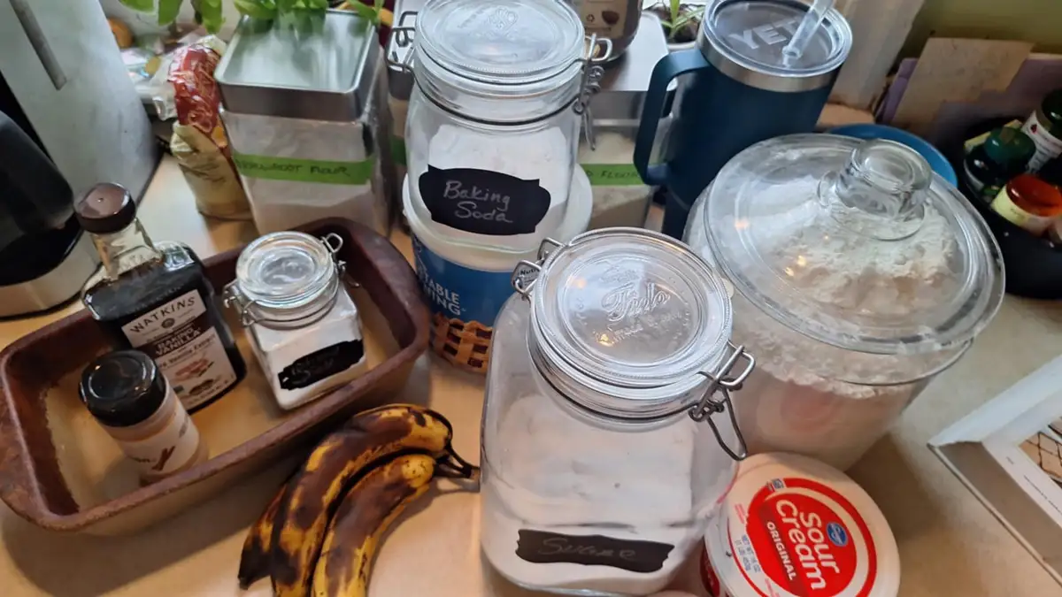 A kitchen counter displays various baking ingredients including jars labeled 'Baking Soda' and 'Sugar', a container of sour…