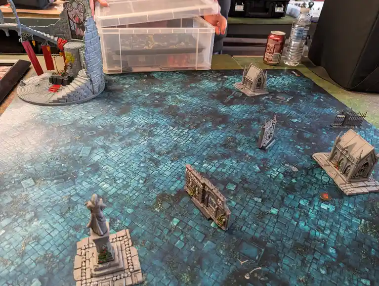 A tabletop miniature wargaming setup with a blue-tiled battlefield, gothic-style terrain pieces, and a hand lifting a clear…
