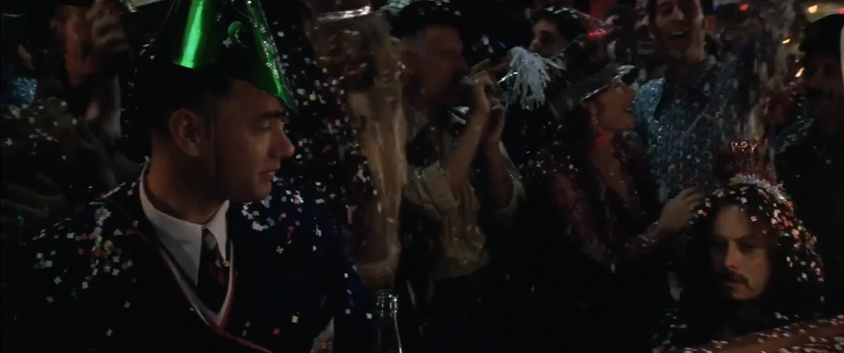 A man in a green party hat and suit covered in confetti looks bored while surrounded by a lively, celebratory crowd at a…