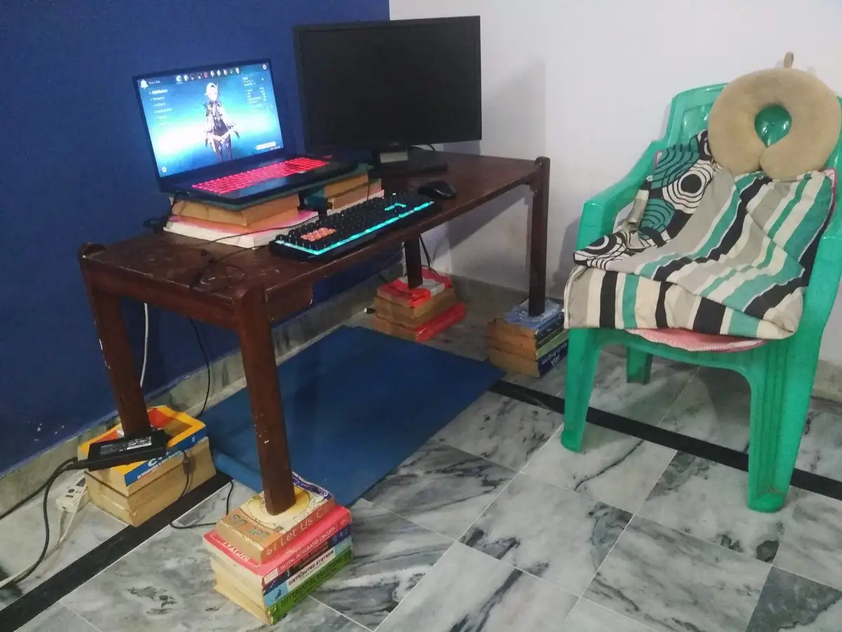 A wooden desk is propped up by stacks of books to elevate a laptop and an external monitor, with a glowing keyboard and…