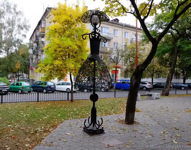A black metal sculpture shaped like a woman in a dress stands in a park, with cars and apartment buildings in the…