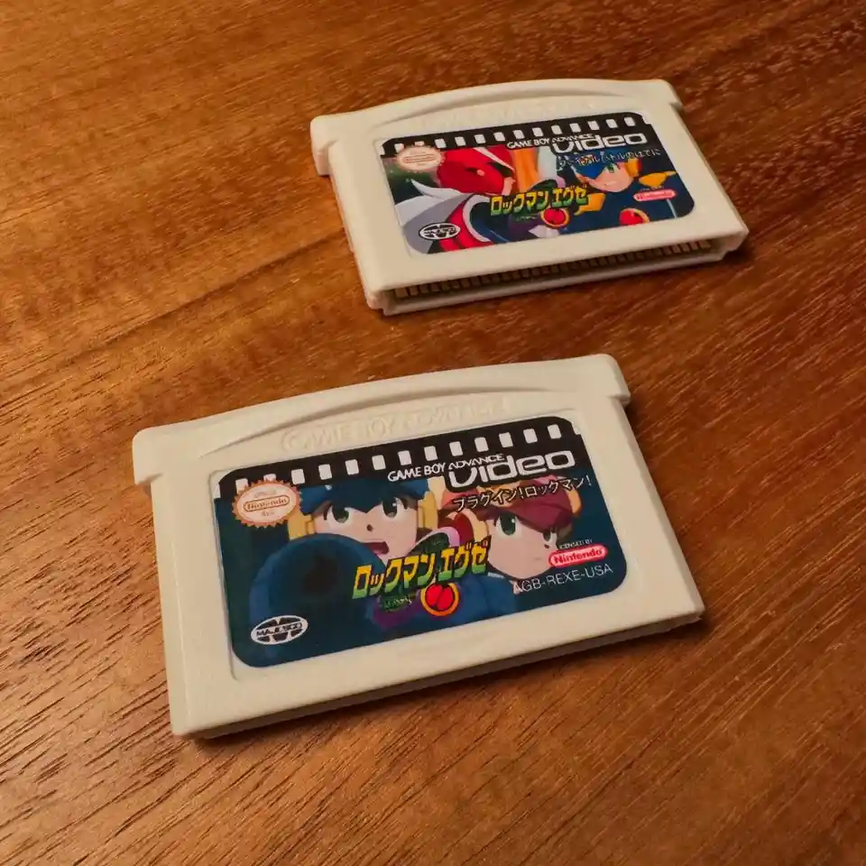 Two Game Boy Advance Video cartridges for the Japanese game 'Rockman EXE' lie on a wooden surface. The top cartridge shows…