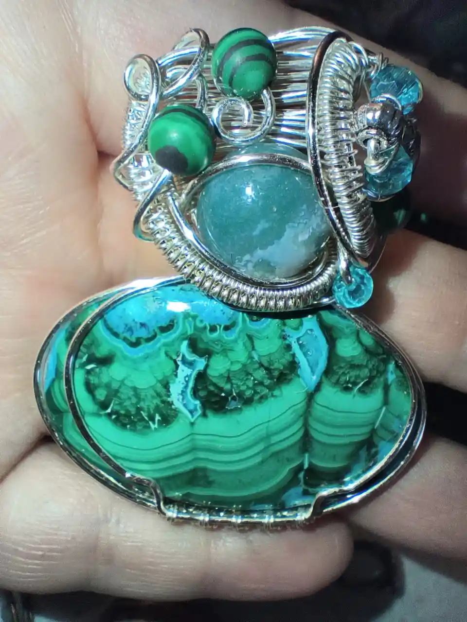 A hand holds a large, ornate pendant made of silver wire wrapping around green malachite and turquoise stones, with smaller…