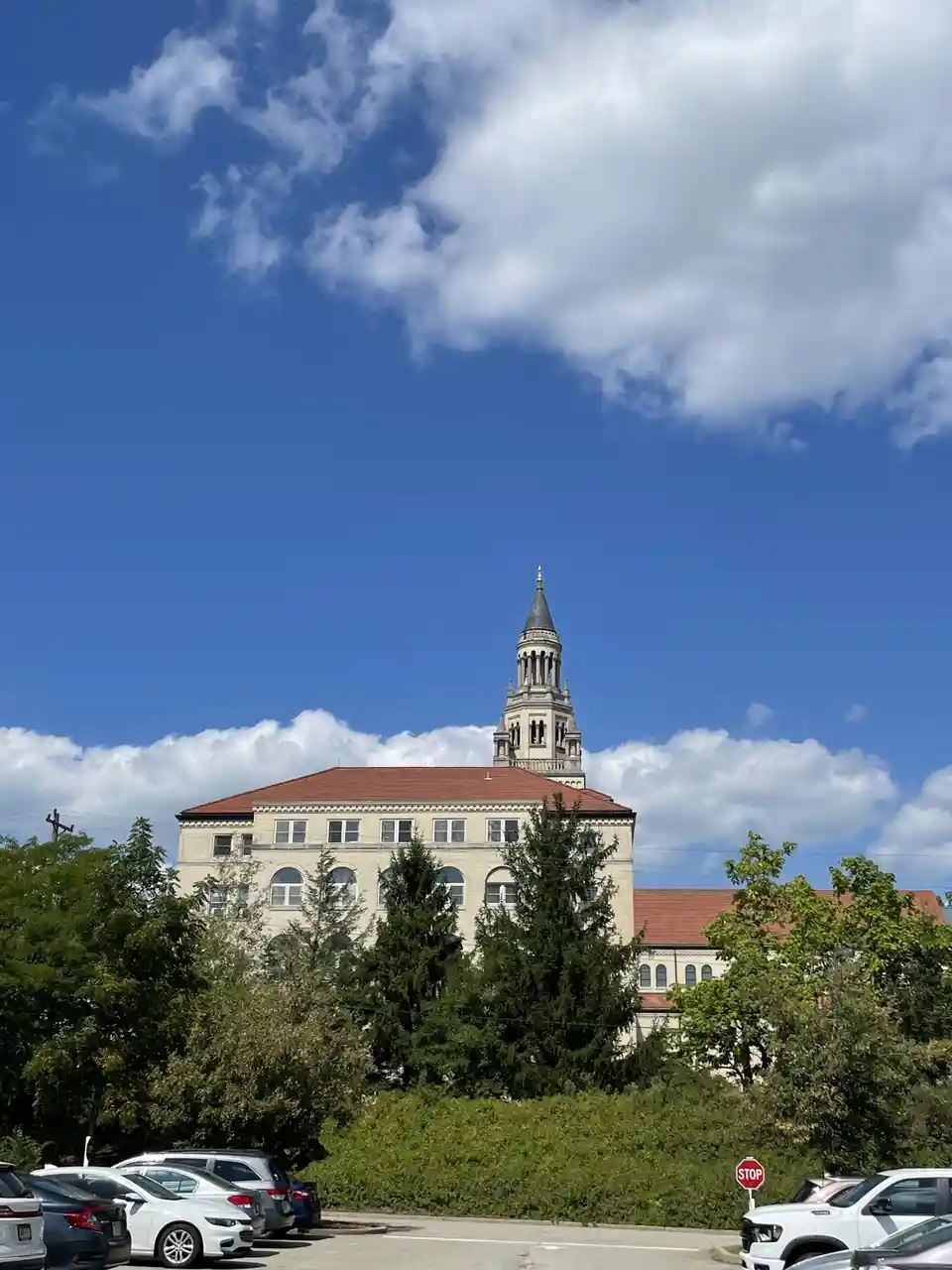 A photograph of a large, light-colored institutional building with a prominent clock tower and red roof, viewed from a…