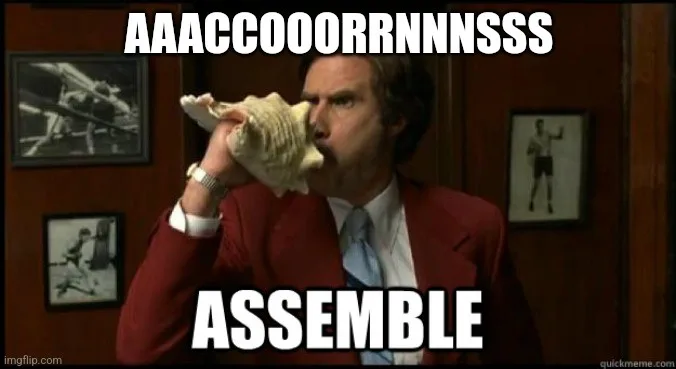 A man in a red suit and blue tie holds a large conch shell to his mouth in an office setting with framed photos on the wall…