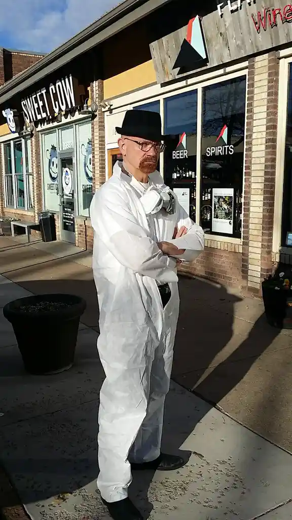 A man in a white hazmat suit, black fedora, and glasses stands with arms crossed outside a store named 'Sweet Cow' that also…