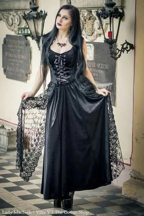 A woman in a black gothic corset dress with lace trim stands in a hallway with ornate wall sconces and plaques resembling…